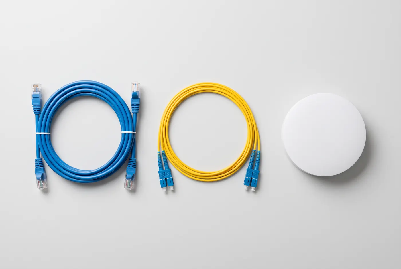 Vue de dessus de trois supports côte à côte : un cordon cuivre bleu à connecteurs RJ45, une jarretière fibre jaune à connecteurs bleus, et une borne Wi-Fi blanche et ronde.